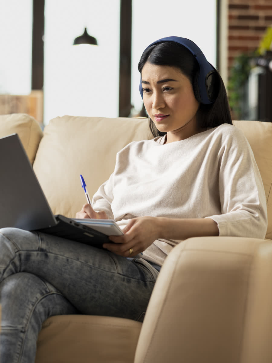 A professional wearing headphones reviews project slides on her laptop from a sofa at home