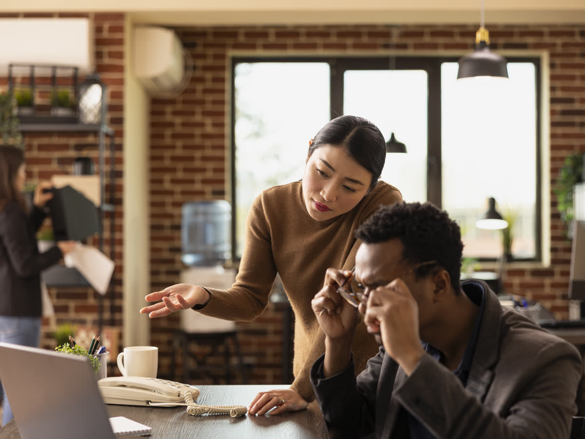 A manager leans over to help a colleague at his desk in a brick-walled office